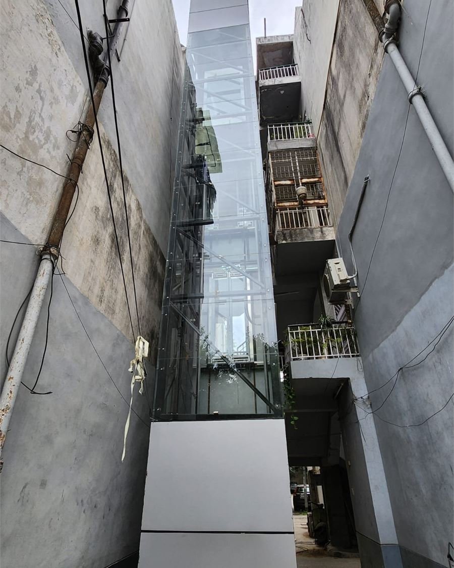 A glass and steel lift shaft built into the narrow gap between two existing buildings, seen looking up from ground level