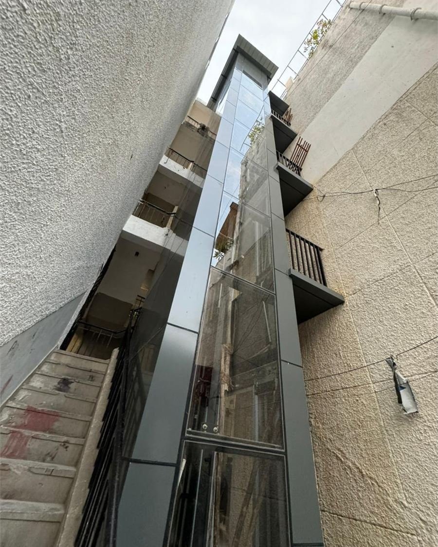 A completed structure lift clad in grey panels and glass, standing in a narrow alley beside a staircase