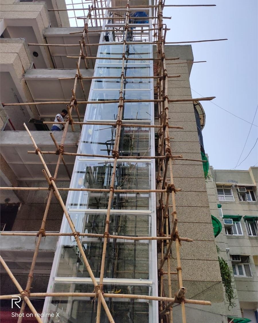 A structure lift under installation against an apartment block, surrounded by bamboo scaffolding with a worker on the balcony